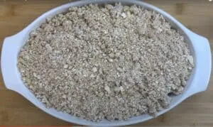 Homemade apple crumble in a white dish on a wooden surface, ready to bake.